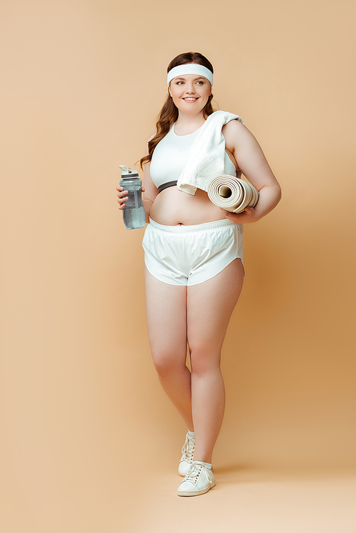 Plus size sportswoman with fitness mat and sports bottle looking away and smiling on beige