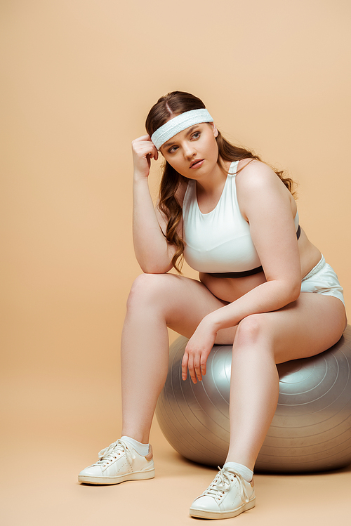 Plus size sportswoman looking away and sitting on fitness ball on beige background