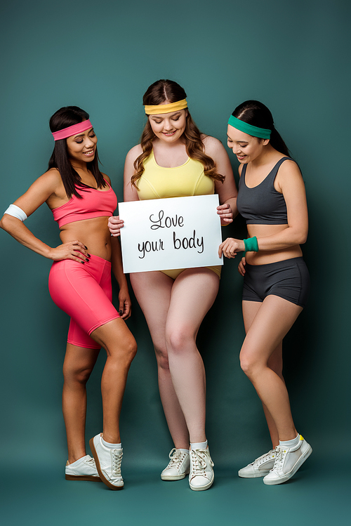 Multiethnic sportswomen smiling and showing placard with love your body lettering on green background