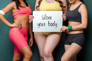 Cropped view of multicultural sportswomen showing placard with love your body lettering on green background