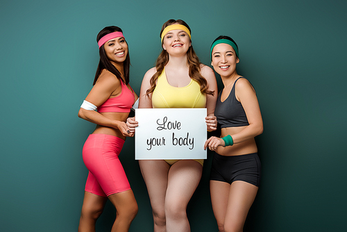 Multicultural sportswomen smiling,  and showing placard with love your body lettering on green background