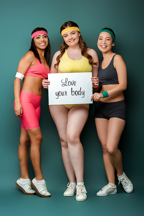Multicultural sportswomen smiling,  and presenting placard with love your body inscription on green