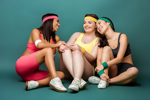 Multiethnic sportswomen sitting with crossed legs, smiling and looking at each other on green