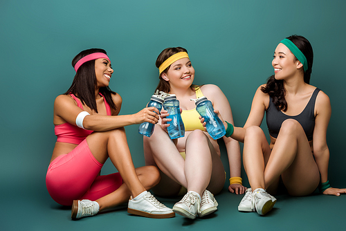 Multiethnic sportswomen sitting with crossed legs, smiling, holding sports bottles and looking at each other on green