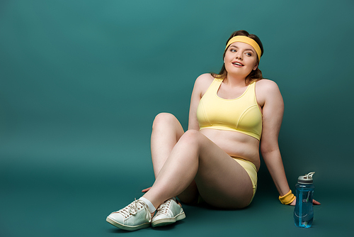 Plus size sportswoman sitting with crossed legs near sports bottle on green background