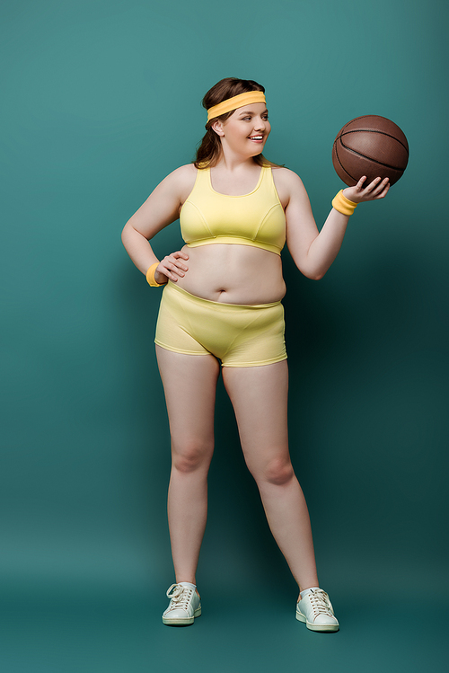 Plus size sportswoman with hand on hip looking at ball and smiling on green background