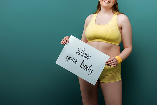 Cropped view of plus size sportswoman smiling and showing placard with love your body lettering on green