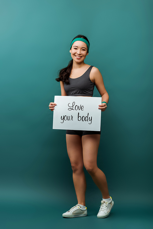 Attractive asian sportswoman smiling and holding placard with love your body lettering on green