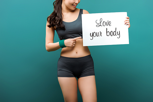 Cropped view of sportswoman smiling and pointing at placard with love your body lettering on green background