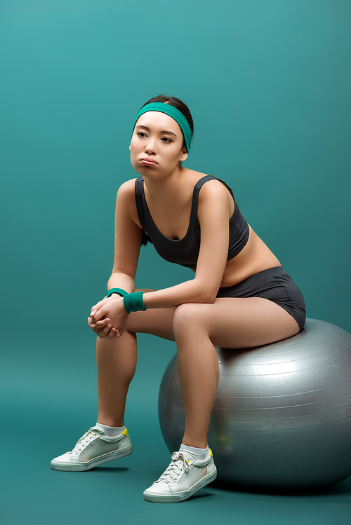 Tired asian sportswoman sitting on fitness ball on green background