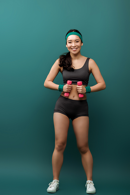 Front view of asian sportswoman , smiling and holding dumbbells on green background