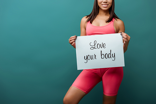 Cropped view of african american sportswoman smiling and holding placard with love your body inscription isolated on green