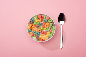 top view of bright colorful breakfast cereal in bowl near spoon on pink background