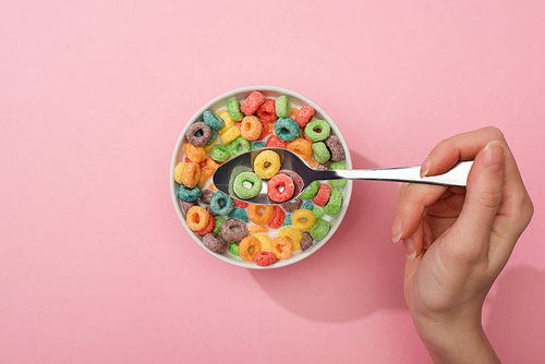 partial view of woman eating bright colorful breakfast cereal from bowl with spoon on pink background