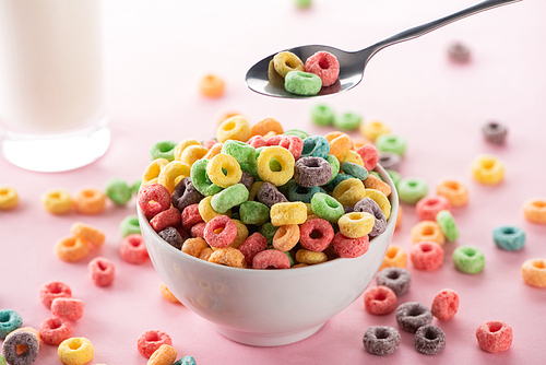 selective focus of bright multicolored breakfast cereal in bowl near glass of milk and spoon on pink background