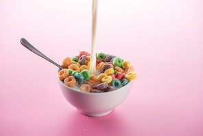 bright multicolored breakfast cereal in bowl with pouring milk and spoon on pink background