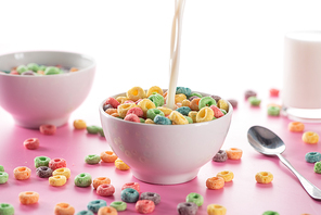 selective focus of bright multicolored breakfast cereal in bowl with pouring milk near spoon on pink background