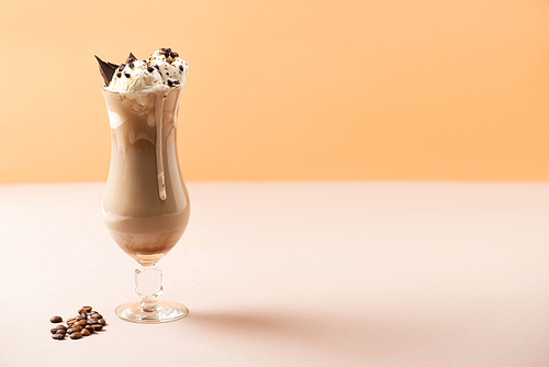 Glass of milkshake with ice cream and chocolate morsels near coffee grains on beige and orange