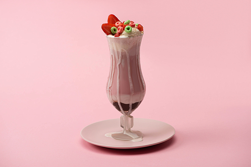 Glass of delicious milkshake with candies and strawberry on plate on pink background
