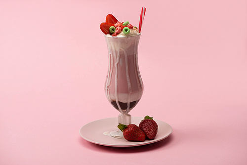 glass of milkshake with  straw, candies and strawberries on plate on pink background