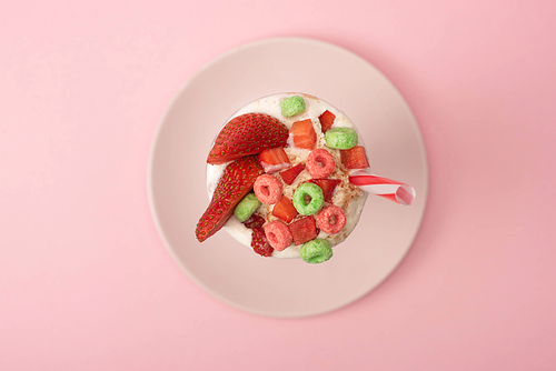 top view of milkshake with  tube, strawberry halves and candies on plate on pink background
