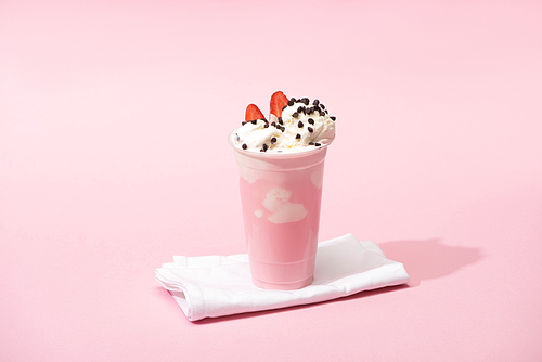Disposable cup of milkshake with chocolate chips and strawberry halves on napkins on pink background