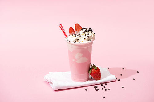 disposable cup of milkshake with  straw, chocolate chips and strawberries on napkins on pink background