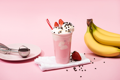 Disposable cup of milkshake with chocolate chips and strawberries on napkins near bananas and plate with scoop on pink