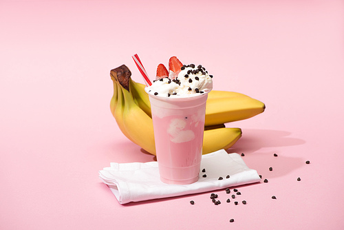 Disposable cup of strawberry milkshake with chocolate chips on napkins near bananas on pink background