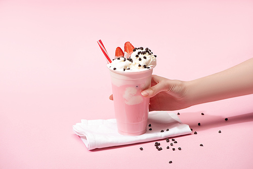 Cropped view of female hand with disposable cup of milkshake with chocolate morsels and strawberry on napkins on pink