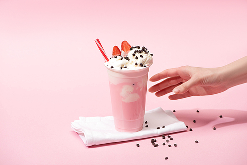 Partial view of female hand with disposable cup of milkshake with chocolate morsels and strawberry on napkins on pink