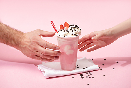 Partial view of female and male hands with disposable cup of strawberry milkshake with chocolate morsels on napkins on pink