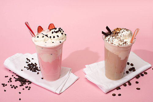Disposable cups of chocolate and strawberry milkshakes with coffee grains on napkins on pink background
