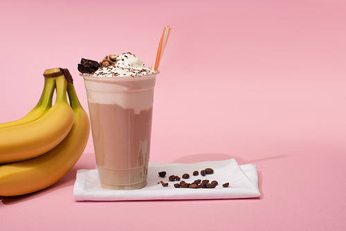 Disposable cup of chocolate milkshake with coffee grains on napkins near bananas on pink