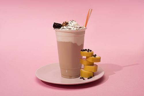 disposable cup of milkshake with  straw, cut banana and chocolate chips on plate on pink background