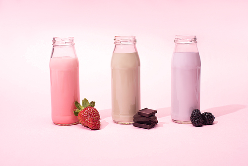 Bottles of milkshakes with strawberry, blackberries and pieces of chocolate on pink background