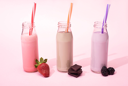 bottles of milkshakes with  straws near strawberry, blackberries and pieces of chocolate on pink
