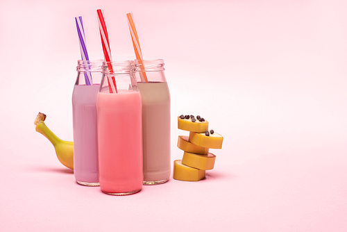 bottles of berry, strawberry and chocolate milkshakes with  straws and cut banana with chocolate chips on pink background