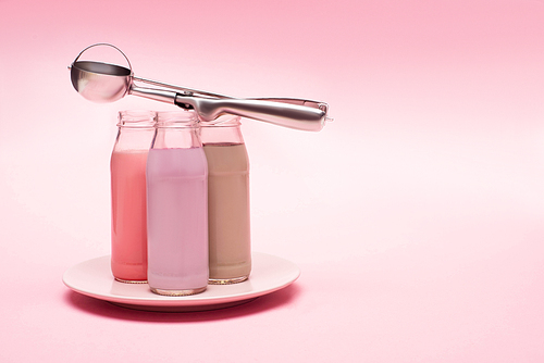 Bottles of berry, strawberry and chocolate milkshakes with scoop on plate on pink background