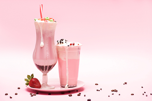 Disposable cup and glass of milkshakes with strawberry on plate and coffee grains on pink background