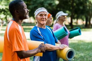 selective focus of happy multicultural senior pensioners standing with fitness mats in park