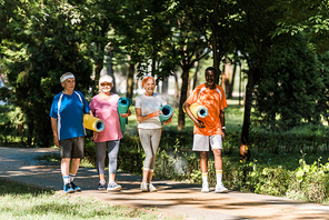 cheerful retired and multicultural pensioners holding fitness mats and walking in park