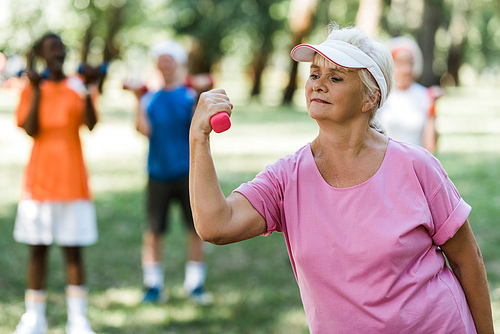 selective focus of senior woman in cap holding dumbbell while exercising near pensioners