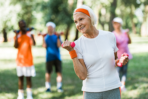 selective focus of cheerful senior woman holding dumbbells while exercising near pensioners