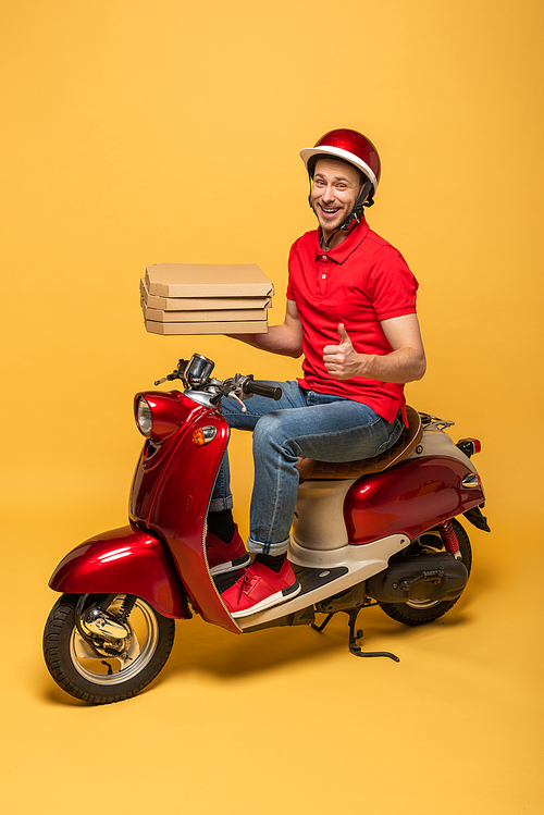smiling delivery man in red uniform holding pizza boxes and showing thumb up on scooter on yellow background