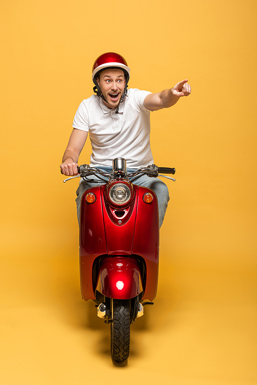 happy delivery man in helmet riding scooter and pointing with finger on yellow background