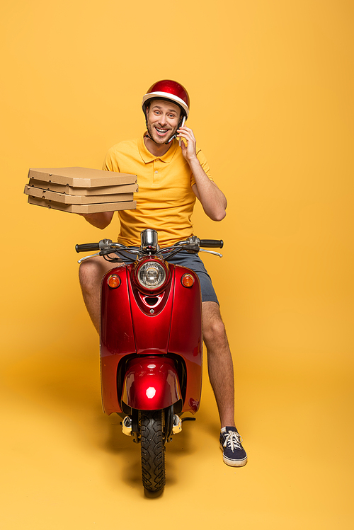 smiling delivery man in yellow uniform riding scooter with pizza boxes and talking on smartphone on yellow background