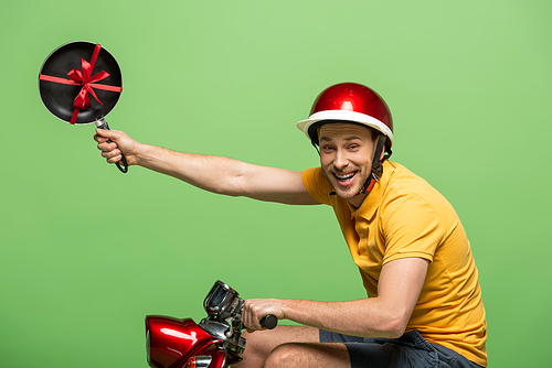 side view of happy delivery man in yellow uniform holding frying pan with bow on scooter isolated on green