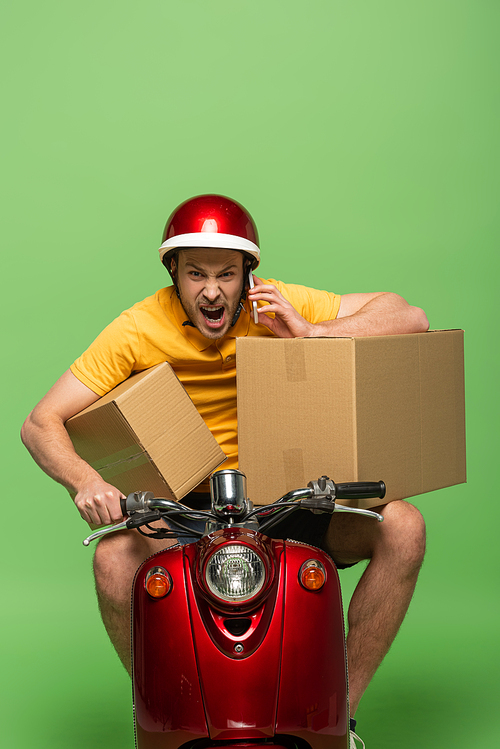 angry delivery man in yellow uniform on scooter with boxes talking on smartphone isolated on green