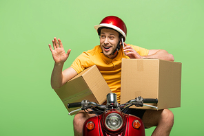 happy delivery man in yellow uniform on scooter with boxes talking on smartphone and waving hand isolated on green